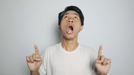 Young Asian man in white shirt shocked while pointing upwards on isolated white background
