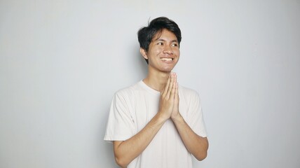 smiling young asian man in white shirt making traditional greeting gesture - obrazy, fototapety, plakaty
