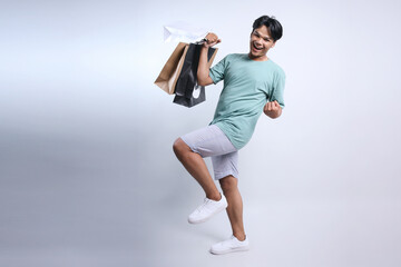 Full length photo of young Asian guy holding shopping bags and celebrate discount sale offer...