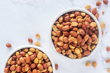 Roasted salted peanuts in white ceramic bowl on white marble background.
