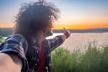 An individual with curly hair points towards a lake at sunset, embodying a sense of peace and contemplation. Curly-Haired Person Enjoying a Sunset by the Lake. High quality photo