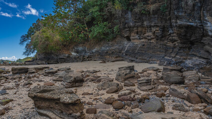 The low tide in the ocean. Exposed layered boulders are scattered on the sand. There is green vegetation on the rocky steep slopes. Clouds in the blue sky. Madagascar. Nosy Be  