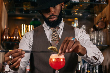 stylish black bartender offering a customer a cocktail decorated with garnish in a traditional...