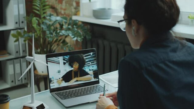 Female renewable energy engineer sitting at office desk and speaking with colleague via video call on laptop when having online business meeting