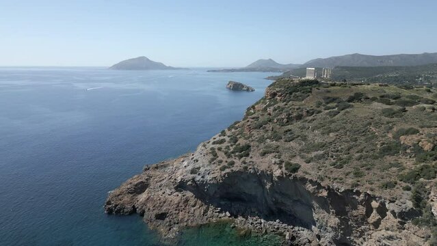 A drone captures the breathtaking view along a cliff in Sunion, Greece, ending with the ancient ruins of the Temple of Poseidon, with an island visible in the distance.