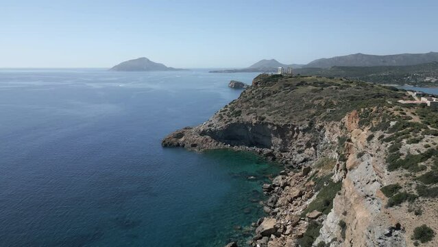A drone captures the breathtaking view along a cliff in Sunion, Greece, ending with the ancient ruins of the Temple of Poseidon, with an island visible in the distance.