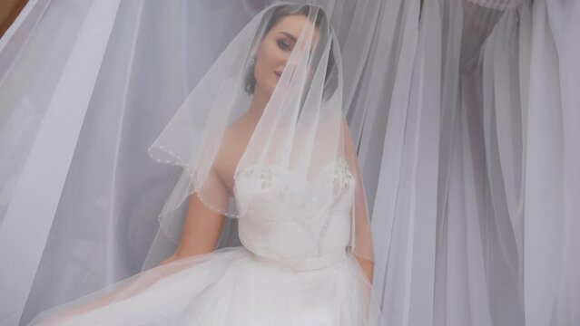 A beautiful young bride in a dress and with a long veil poses in the studio on a white background, she waves her long skirt in front of the camera. The wedding day.