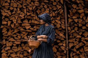 A pioneer woman in a dark blue dress and bonnet standing with a basket of flowers against a wood...