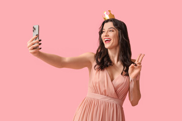 Beautiful young woman in stylish prom dress and paper crown taking selfie on pink background
