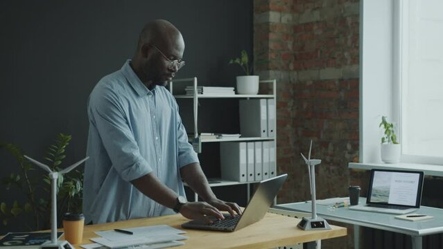 African American engineer standing by desk in modern office and working on laptop when developing green energy project. Medium full shot, copy space