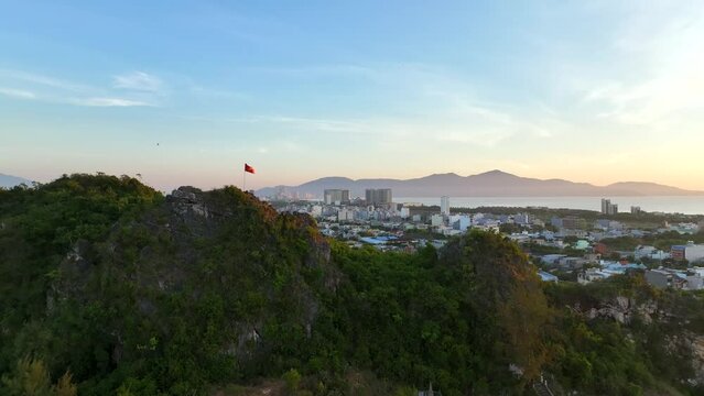 Wide establishing shot of Vietnam flying past Vietnamese flying. Drone video rising up over hill with a Vietnamese flag on it to reveal the city of Da Nang. 