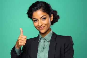 A young Hispanic businesswoman in her early 30s, against a gentle mint green background, showing a thumbs-up gesture with her hand.