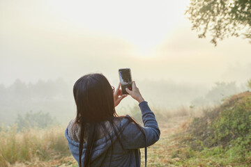 Rear view of woman holding smartphone and taking misty morning - Powered by Adobe