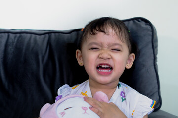 Happy cute toddler sitting on sofa, holding doll, facing camera in living room.