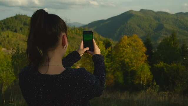 Young woman standing on top of mountain peak photographing scenic landscape using mobile phone device. Happy girl making photos of mountains on cellphone camera