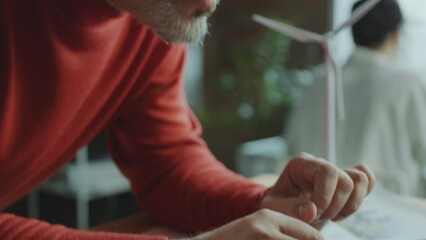 Mature renewable energy engineer with gray hair working on laptop at desk with windmill turbine model during day in the office. Close-up shot