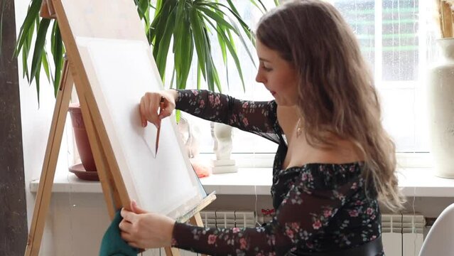 Beautiful woman artist painting with pencil on paper and wooden easel. Female working in art studio in process. Blonde girl with long hair wearing black floral dress
