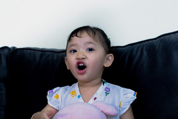 Happy cute toddler sitting on sofa, holding doll, facing camera in living room.