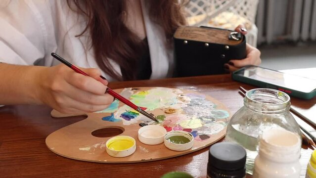 Female artist mixing acrylic colors on palette in studio. Beautiful young woman starts to paint at the table with art tools. Longhaired brunette girl in creative process