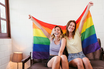 Portraits of happy Caucasian lesbian couple sitting and embracing on a sofa and raising Rainbow flags together in living room. LGBTQ friendship or couples relationships spend quality time together. - 773773050