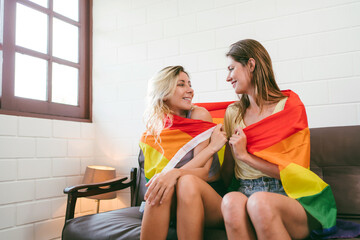 Portraits of happy Caucasian lesbian couple sitting and embracing on a sofa and raising Rainbow flags together in living room. LGBTQ friendship or couples relationships spend quality time together. - 773773062