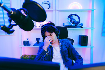 A young Asian man professional gamer wearing jeans jacket sits on a chair with hands over his head, feeling angry and sad when losing the esports competition. Concept of pain from gaming and esports. - 773773691