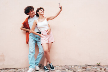 Young smiling beautiful woman and her handsome boyfriend in casual summer clothes. Happy cheerful family. Female having fun. Couple posing in street at sunny day. Near  wall. Take selfie photos