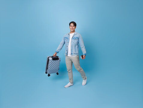 Happy smiling young Asian tourist man holding passport ticket and luggage going to travel on summer holidays isolated on blue background.