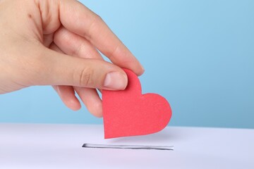 Woman putting red heart into slot of donation box against light blue background, closeup