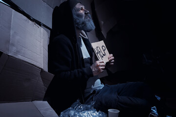 A homeless bearded man sits on boxes on the street and asks for help. Need a homeless person asks for money for food and overnight. - obrazy, fototapety, plakaty