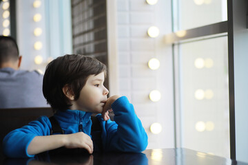A young boy sits at a table near the window in a cafe and waits for a waiter to come up and take an...