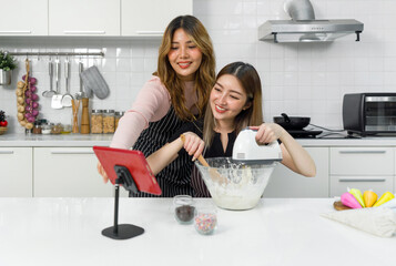 Two women in an apron, smiling and baking together in a kitchen with a tablet, using an electric mixer and a mixing bowl.