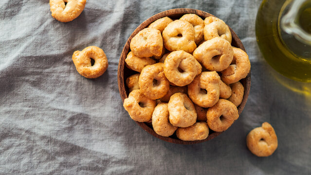 Traditional italian snack taralli or tarallini in wooden bowl over gray linen napkin background. Rustic shot of taralli appetizer with copy space. Top view or flat lay. Horizontal banner