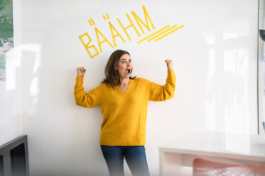 Creative businesswoman flexing muscles in front of white wall with text in office