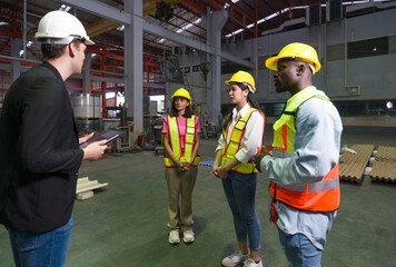 Group of engineer in safety vest meeting with young manager in black suit in an industrial or factory setting. Everyone wearing protective hardhat. Atmosphere in manufactory with large machinery.