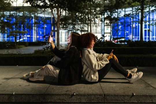 Happy friends sitting back to back and using smart phones at night