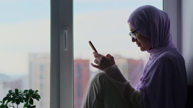 Adult muslim woman in hijab using smartphone sitting on the windowsill at home. Portrait of confidence female in headscarf tapping and scrolling on mobile phone.