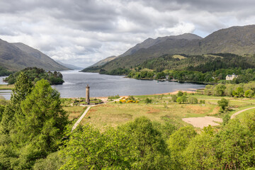The Glenfinnan Monument gracefully punctuates the shores of Loch Shiel, surrounded by the lush, undulating landscape of the Highlands, with the loch stretching into the distance