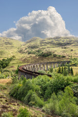 Naklejka premium The historic Glenfinnan Viaduct arcs through the verdant Scottish landscape, its ancient stones contrasting the vibrant natural tapestry under a vast, cloud-filled sky