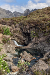 A crisp waterfall spills into the tranquil Fairy Pools of the Isle of Skye, nestled within a rugged landscape with the majestic Cuillin peaks in the backdrop