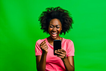 Exuberant woman with afro hair celebrating a victory or good news on her smartphone against a green background - obrazy, fototapety, plakaty