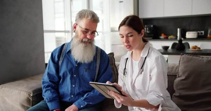 A brunette woman doctor in a white coat holds an electronic tablet in her hands and shows an elderly man in glasses with a lush gray beard what he needs to do to maintain his health in a modern