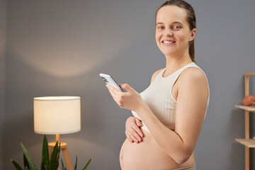 Smiling pregnant woman wearing beige top stroking her bare tummy holding cell phone browsing web...