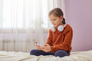 Adorable little girl with brown hair sitting on bed using mobile phone wearing headphone over neck...