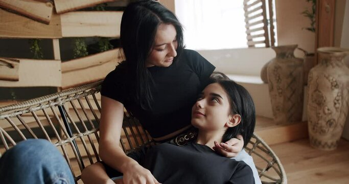Friendship concept. LGBT rights, Lesbian family. Low angle handheld shot of young lesbian couple smiling and touching hands gently while sitting on sofa against window in light living room at home.