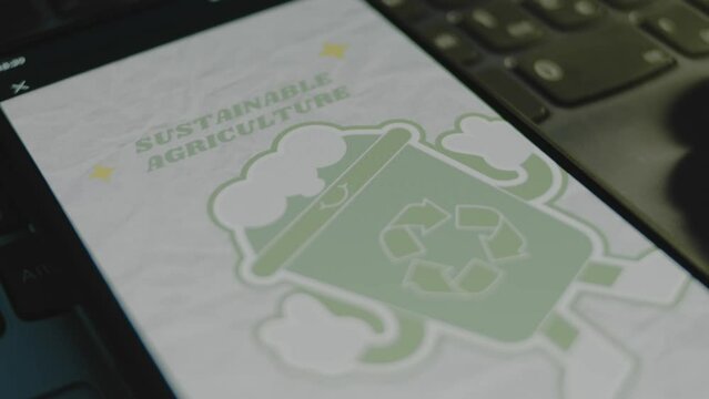 Sustainable agriculture inscription on smartphone screen. Green friendly plastic cup image with a recycle sign on grey background. Environment concept. Male hand flapping fingers cheerfully