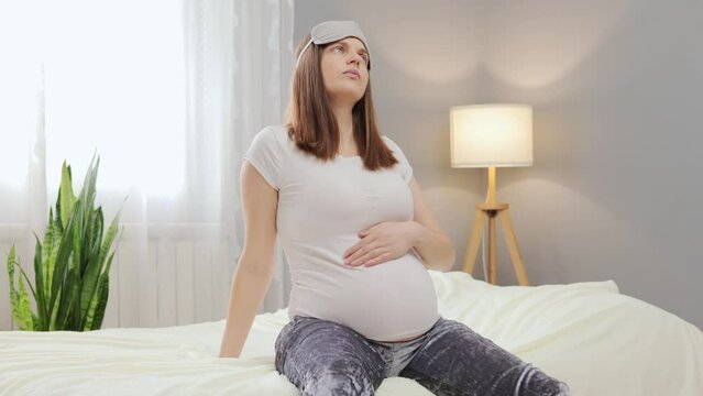 Young beautiful pregnant woman feeling pain with labor contraction sitting on bed in bedroom preparing to give a birth looks nervous stroking her belly