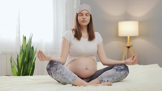Relaxed pregnant woman doing yoga while sitting on bed in lotus pose trying to calm down wearing pajama and sleeping mask starting her morning with meditation