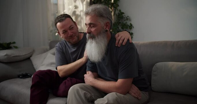 A sad man with gray hair and a lush beard with a black earring in his ear and in a gray T-shirt is sad and his middle-aged brunette boyfriend supports him Sitting on the sofa in a modern apartment in