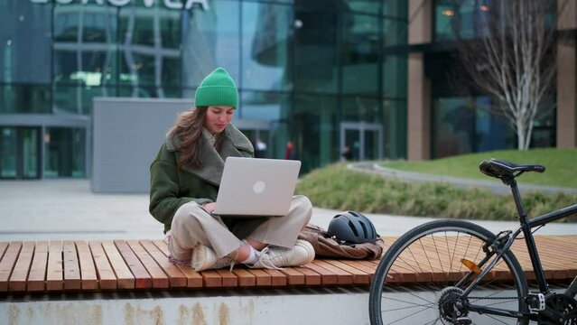 University student on bench in front of university building working on laptop. Fashionable Gen z woman traveling to school by bike. City commuter student, sustainable lifestyle. - Powered by Adobe